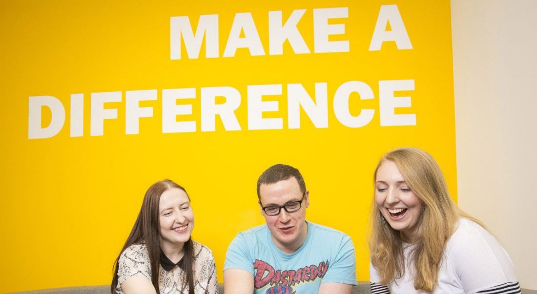 Three young professionals are smiling and interacting, sitting in front of a yellow wall with white writing that says 'make a difference'.