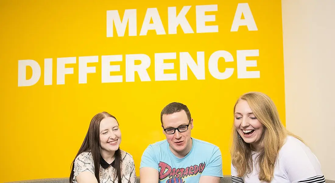 Three young professionals are smiling and interacting, sitting in front of a yellow wall with white writing that says 'make a difference'.