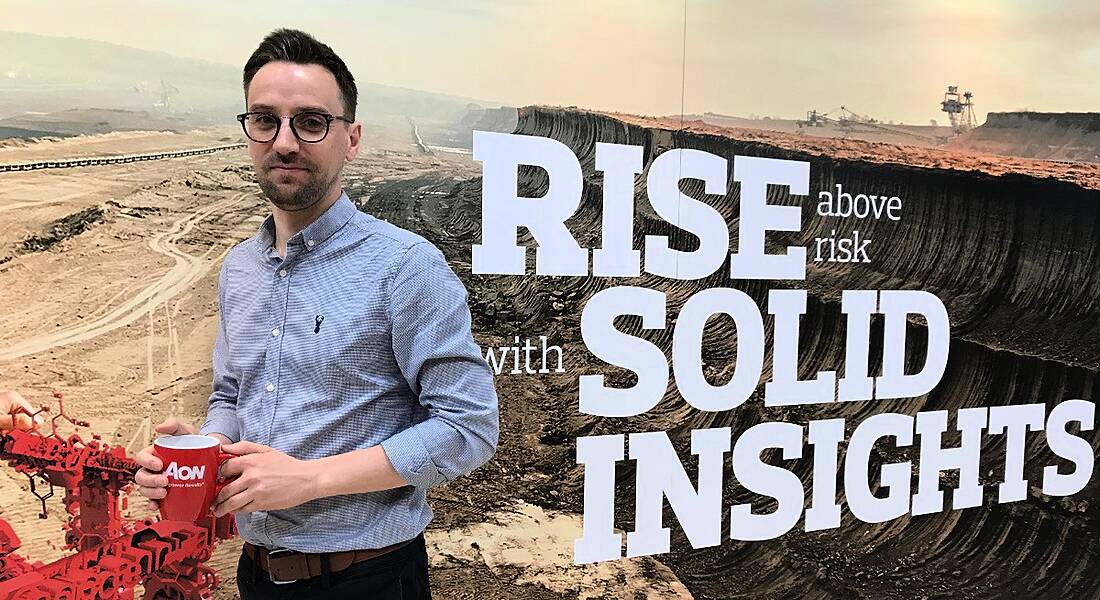 A young professional man is standing against a wall decorated with mountain imagery reading 'rise above risk with solid insights', looking into the camera and holding a red Aon-branded cup.