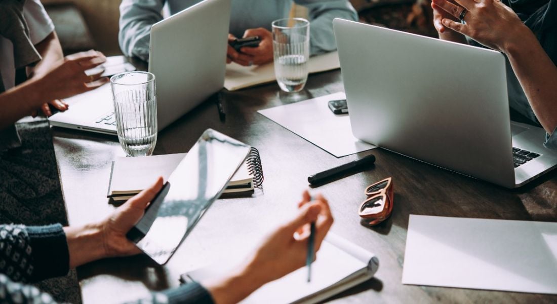 Laptop, mobile phone, tablet and documents on a working table in an office setting, with colleagues sitting around it.