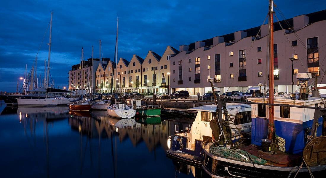 The harbour of Galway city at night with boats docked in front of a row of buildings.