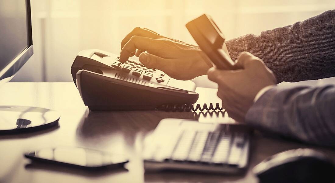 Hand using a phone in the office, with a keyboard, mouse, mobile, and monitor also on the desk.