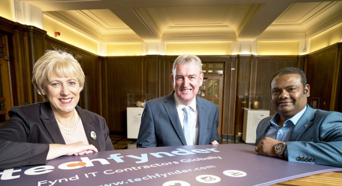 Two men and a woman in a regal-looking room huddled around a banner that says Techfynder while wearing business attire.