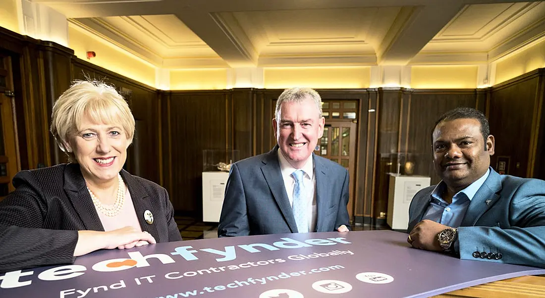 Two men and a woman in a regal-looking room huddled around a banner that says Techfynder while wearing business attire.