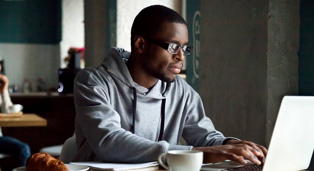 A man in casual clothes in a café setting, representing remote working on a laptop.