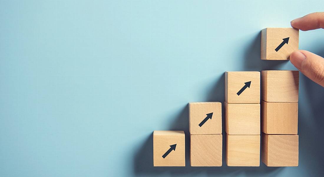 Wooden building blocks being placed on top of one another by a hand, with arrows pointing upwards against a blue background.