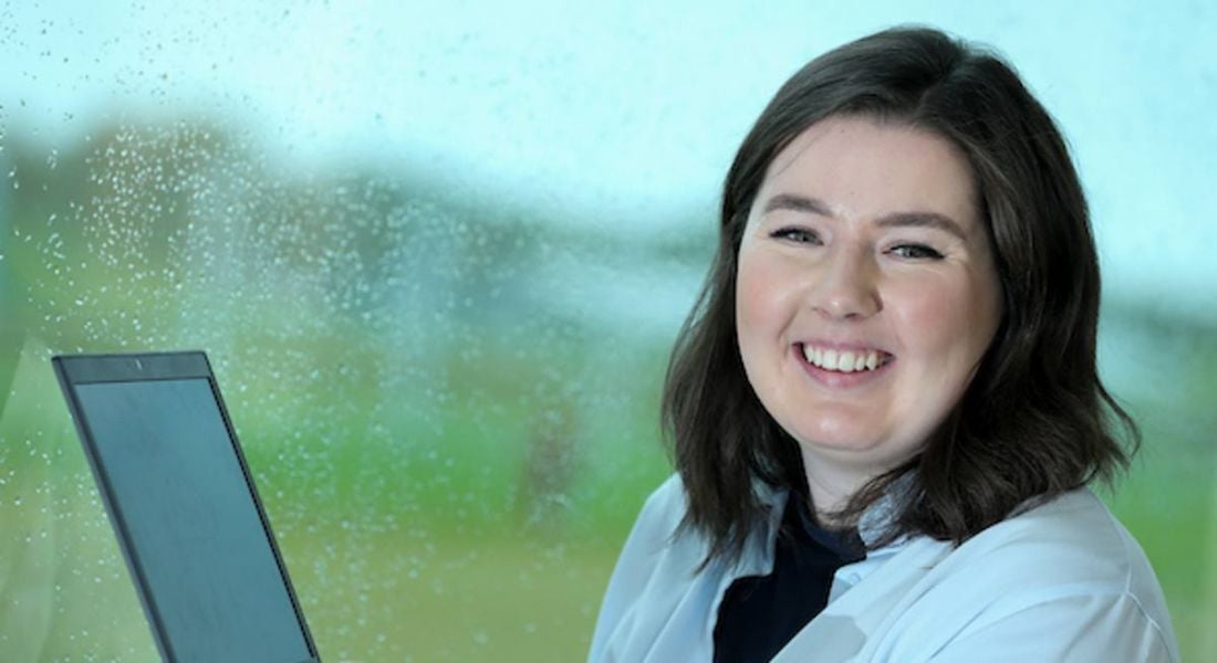 A young professional woman in a lab coat is smiling at the camera with her back to a window, holding a laptop up.