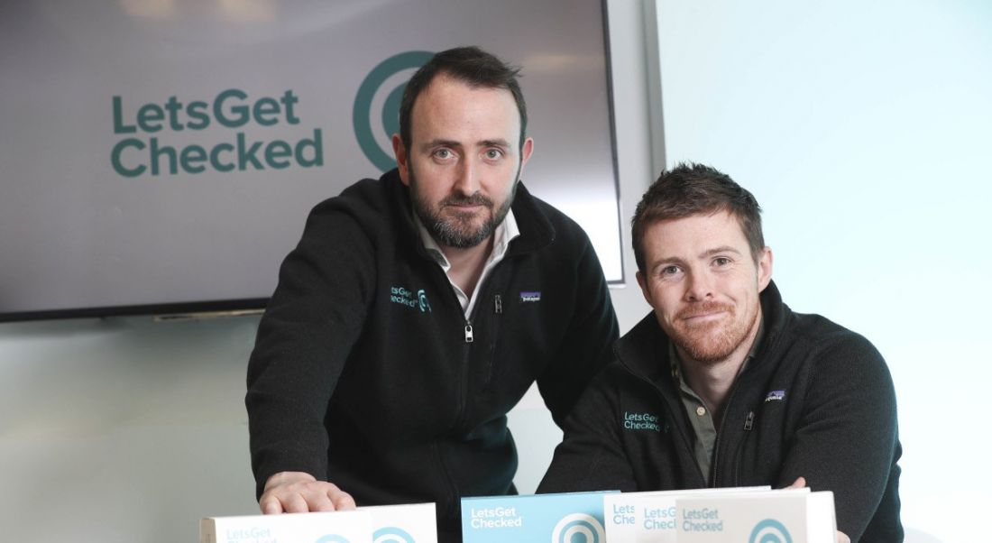 Two young men in black sweaters sitting at a table with LetsGetChecked turquoise logo in the background.