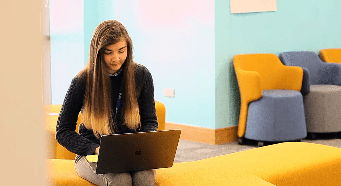 A young woman is sitting on a colourful yellow couch and working on her laptop in Liberty IT.