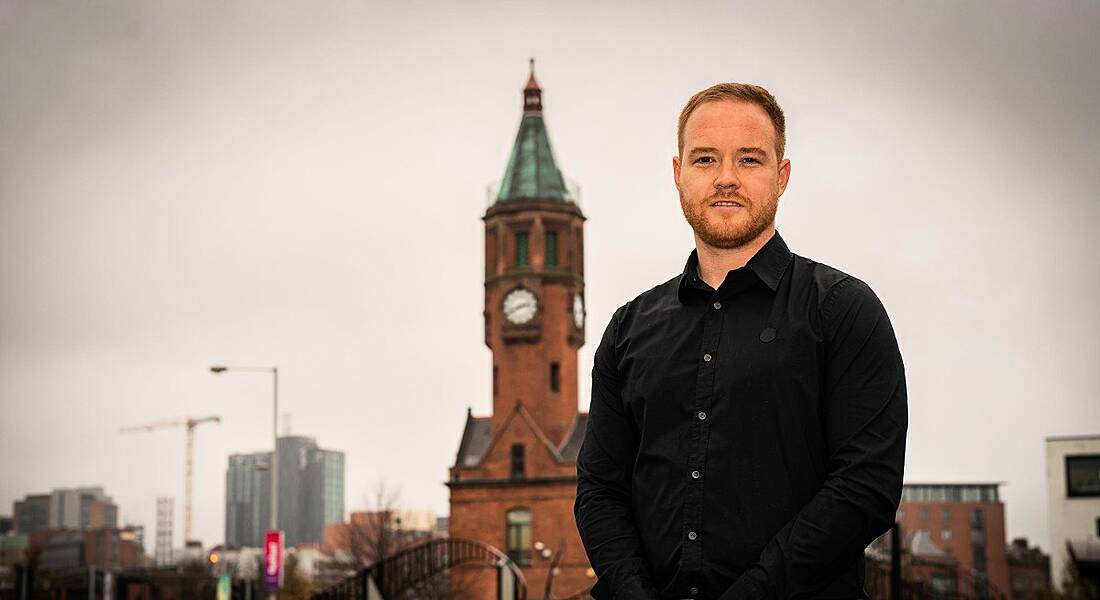 A young man in dark shirt posing against backdrop of a clocktower on overcast day.