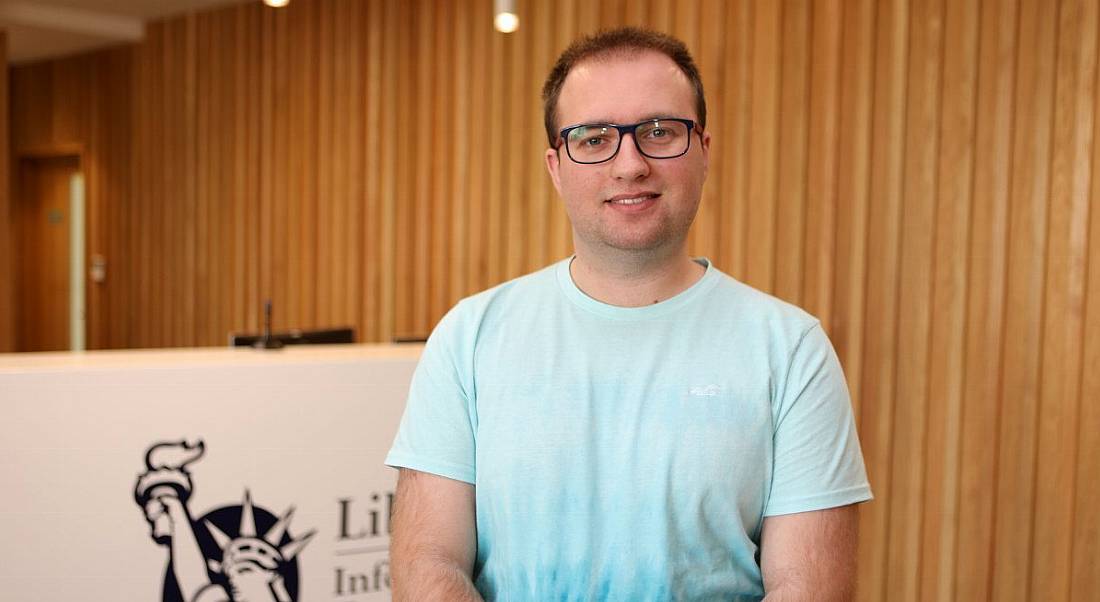 A young professional man in casual clothing is smiling into the camera at the reception area in Liberty IT.