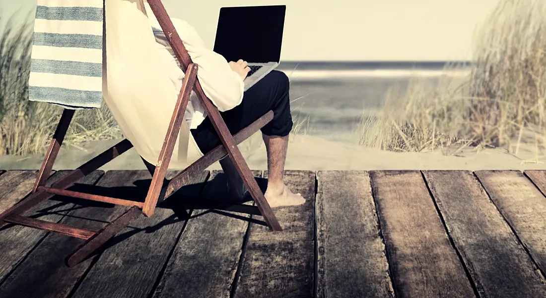 Person sitting in a deckchair on decking at a beach, wearing a white sweater and working on a laptop.