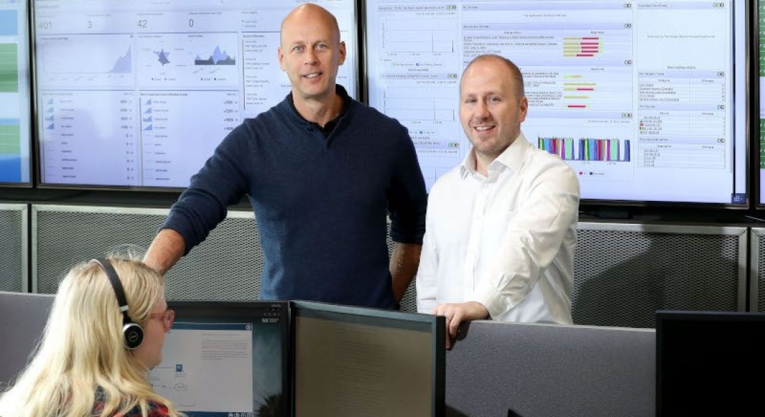 Two men stand in an office full of computers in front of a graphics-covered screen, while a woman with blonde hair works at a desk.