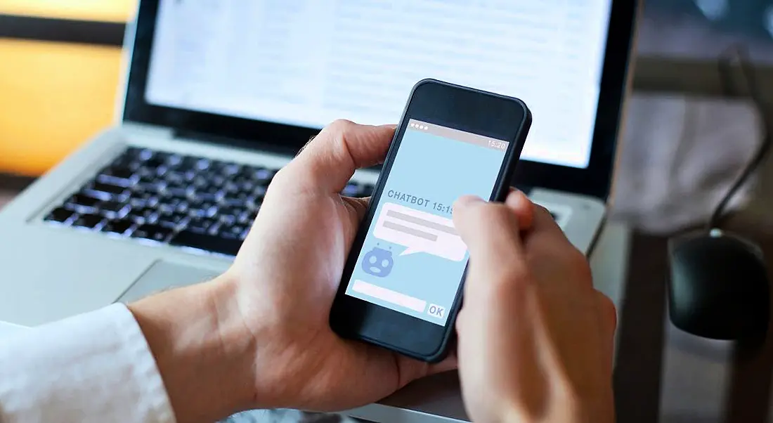 Hand holding a phone an interacting with a chatbot with a laptop in the background.