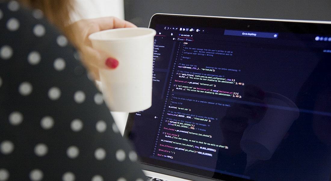 Woman in a spotted top holding a coffee cup and working on code at a computer.