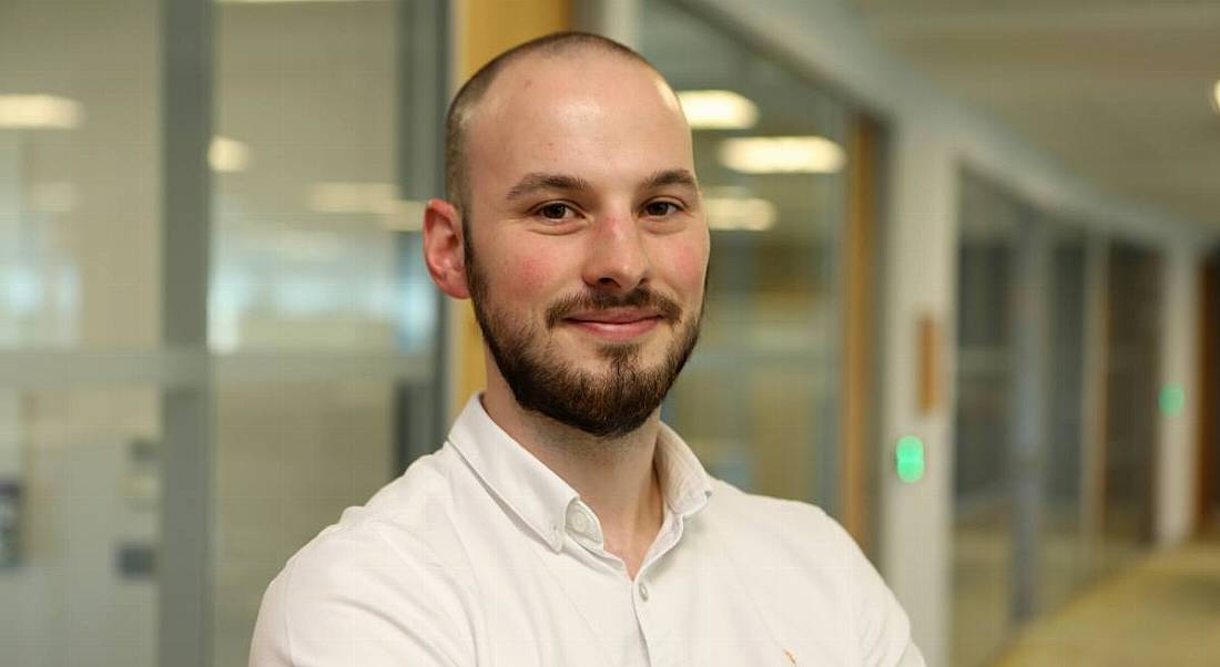 Headshot of a professional young man smiling into the camera in a white shirt, while standing in a corporate setting.