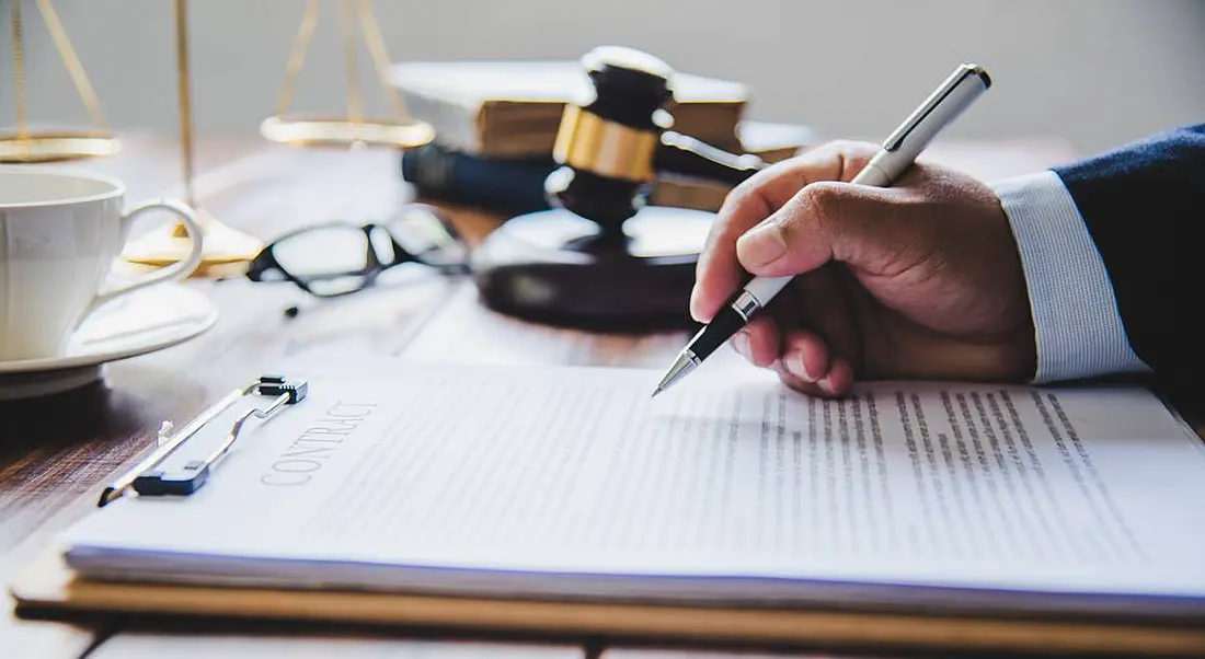 Mallet of a judge on a wooden table, beside a hand that is holding a pen and writing on a document.