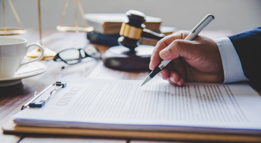 Mallet of a judge on a wooden table, beside a hand that is holding a pen and writing on a document.