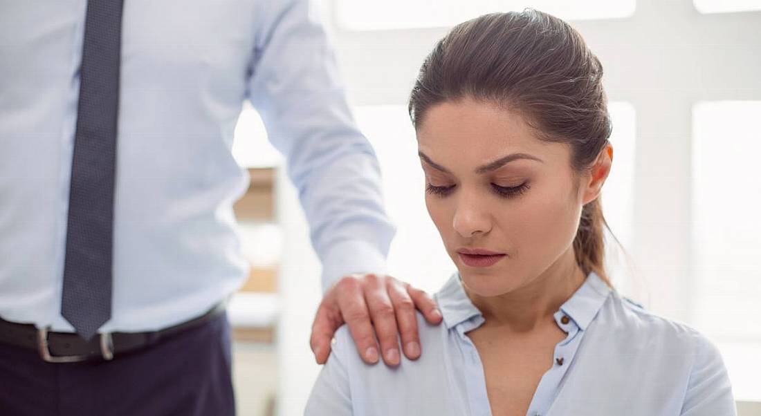 A professional woman sitting down is looking uncomfortable as a male colleague places his hand on her shoulder, symbolising sexual harassment.