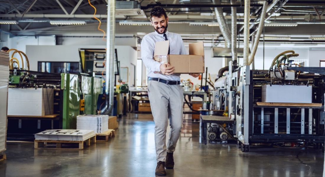 Smiling employee walking away from desks in a modern office and carrying a box of his belongings.