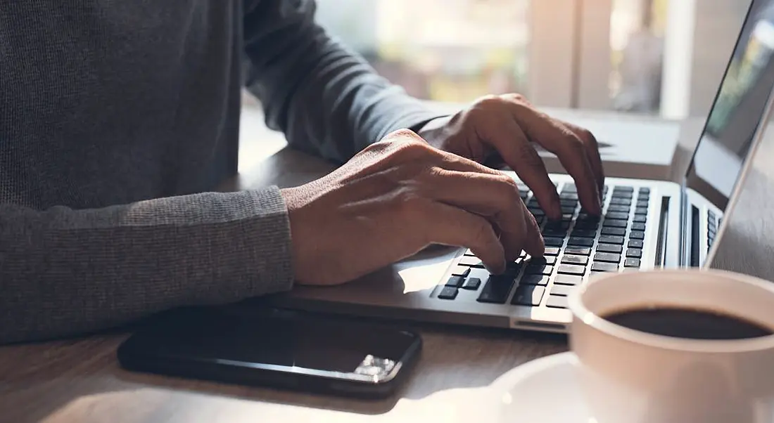 Hands typing on a laptop keyboard with mobile smartphone on office desk at home, working freelance.