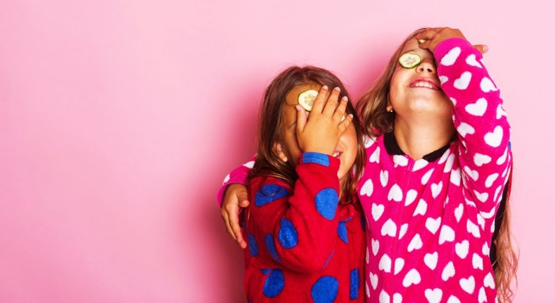 Girls in colourful polka-dotted pyjamas hugging and smiling with cucumber slices on their eyes.