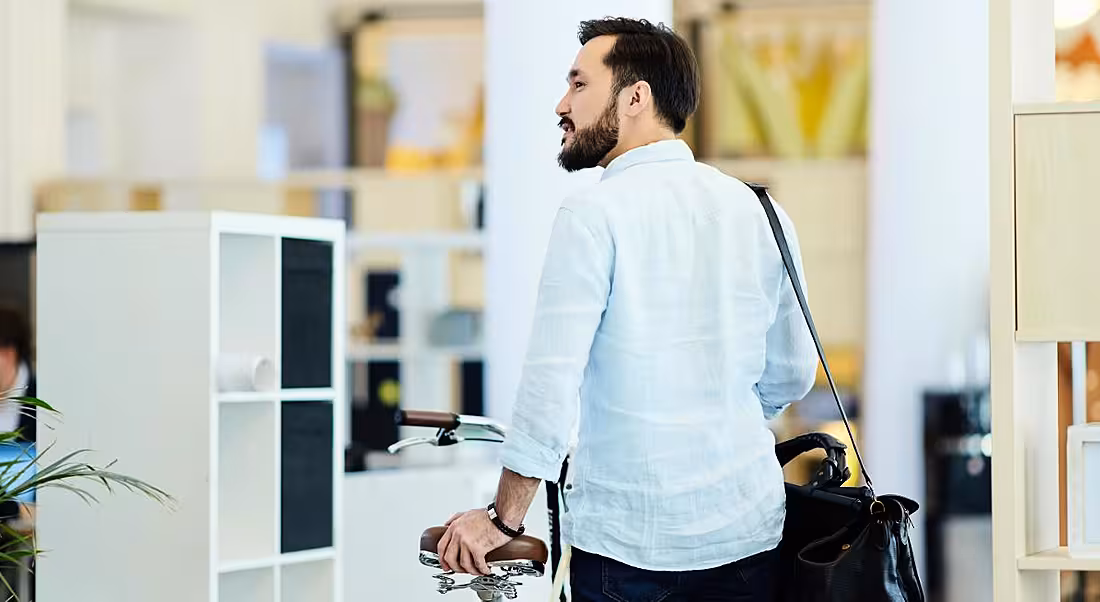 A professional man in casual clothing has his back to the camera but is looking back and smiling at coworkers while leaving a modern office with his bike.