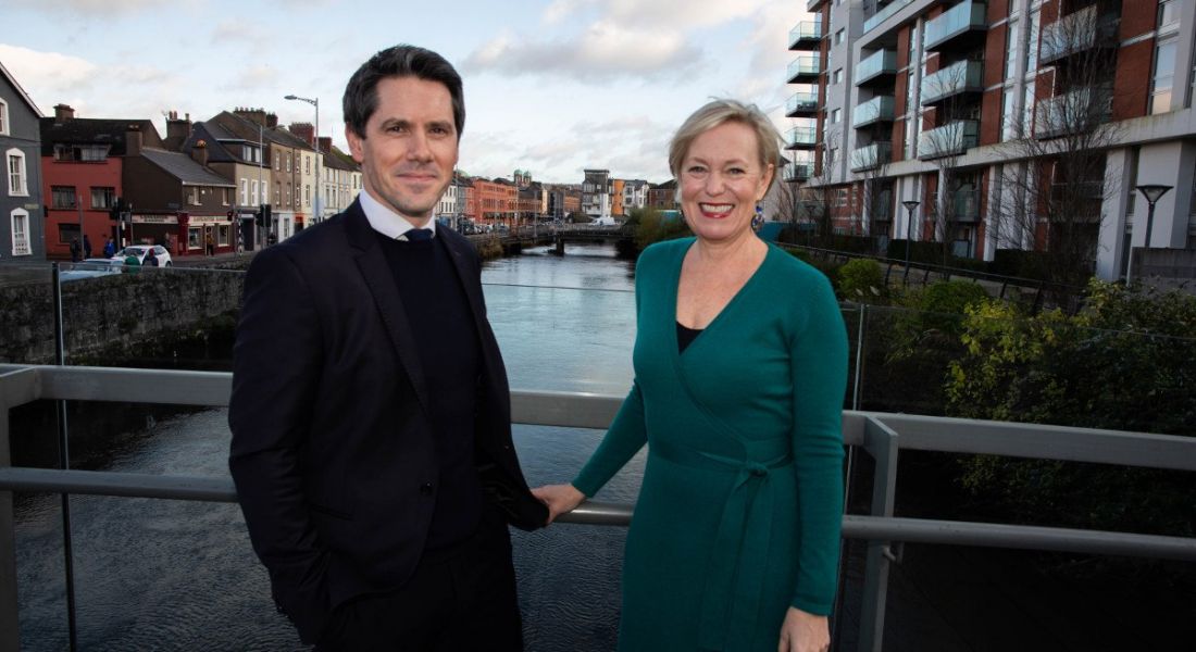 A professional man and woman are standing outside in front of the river in Cork, smiling into the camera.