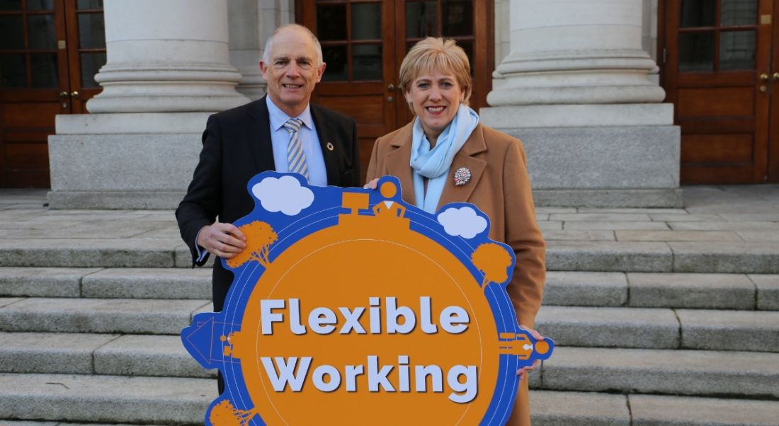 A man and woman are standing outside a government building on steps, holding a bright sign reading 'flexible working'.