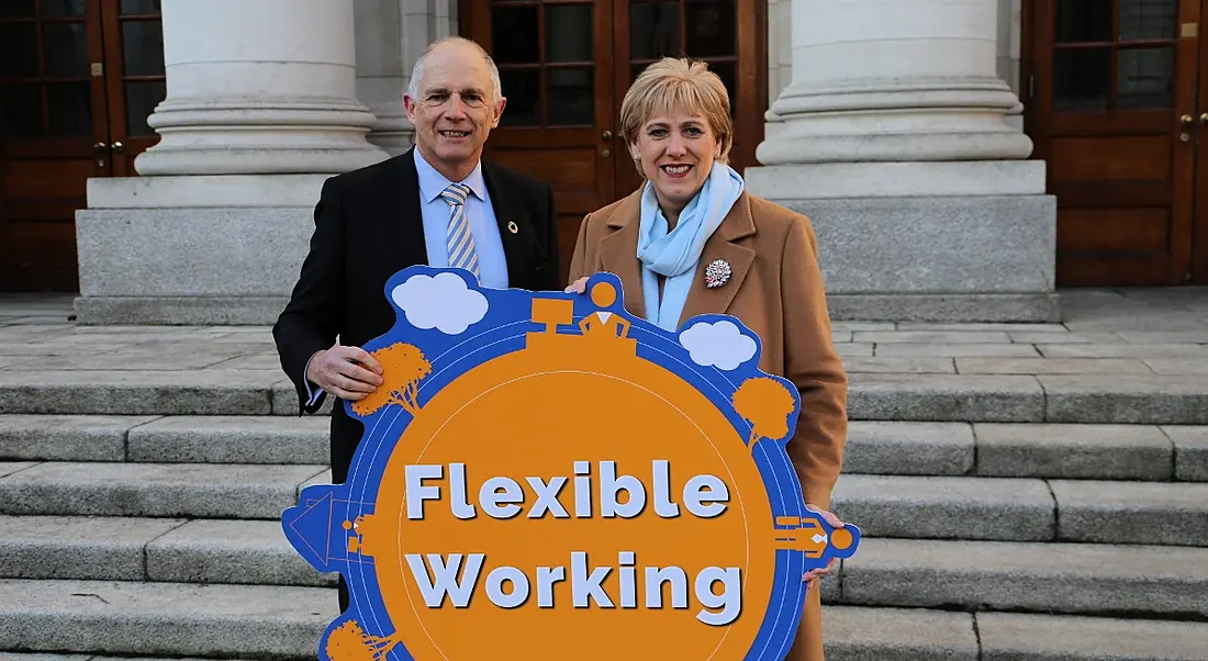 A man and woman are standing outside a government building on steps, holding a bright sign reading 'flexible working'.