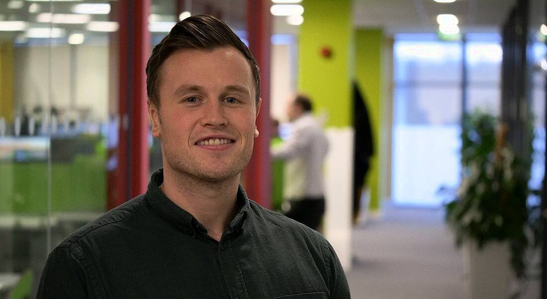 A young professional man is standing in a hallway at Genomics Medicine Ireland, smiling into the camera.