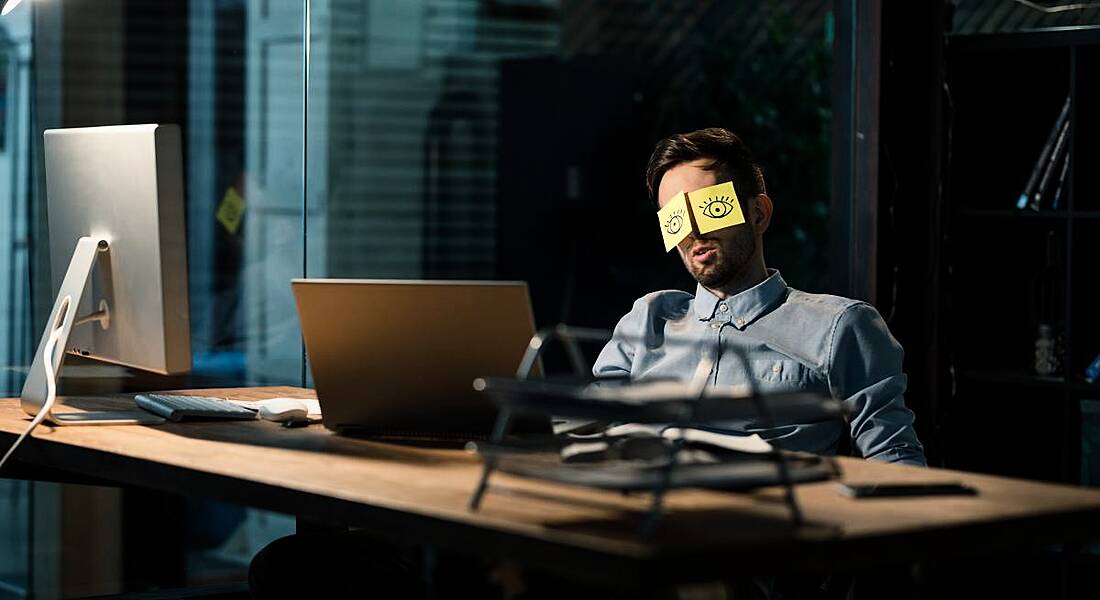 A man sleeping in a dark office, sitting at a table with his eyes covered by stickers that have eyes drawn onto them.