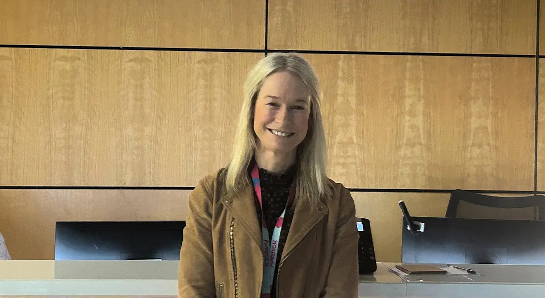 A woman wearing a tan-coloured jacket smiles in front of a reception desk. She is Nollaig Murphy, a HR business partner at Merck.