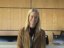 A woman with shoulder-length straight brown hair and a white shirt smiles at the camera in front of a cream-coloured wall.