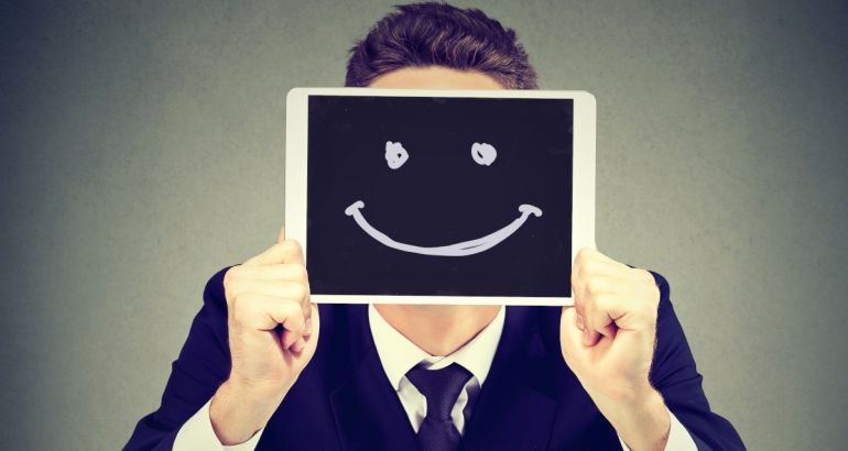 A man holds up a tablet bearing an unnatural smiling face, to represent toxic positivity in the workplace.