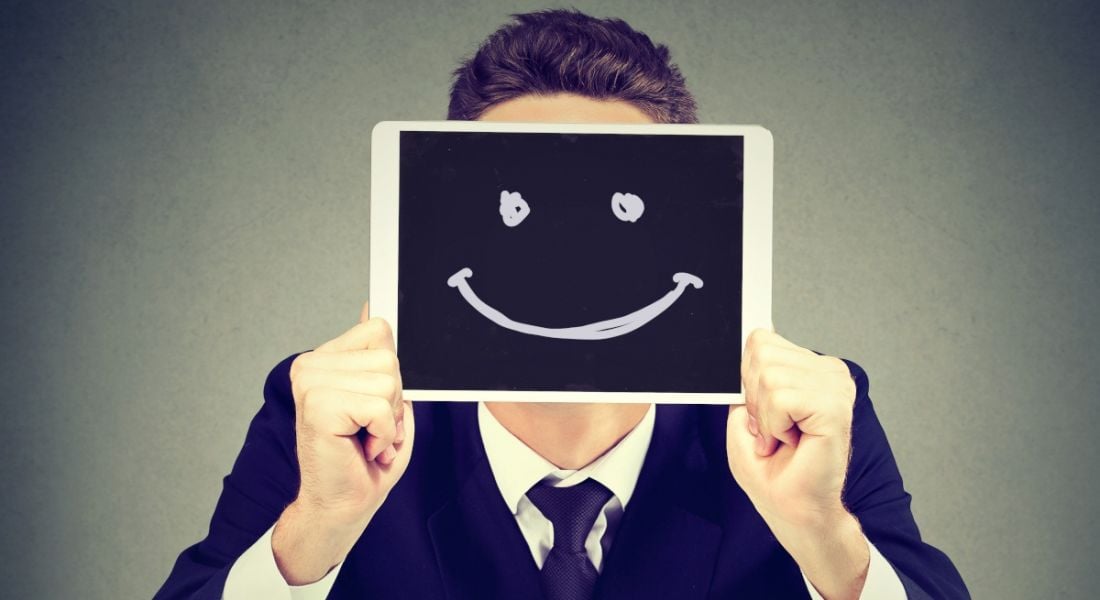 A man holds up a tablet bearing an unnatural smiling face, to represent toxic positivity in the workplace.