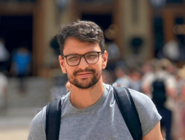 A man with a beard wearing glasses and a backpack smiles at the camera in front of a busy street. He is Paulo da Costa.