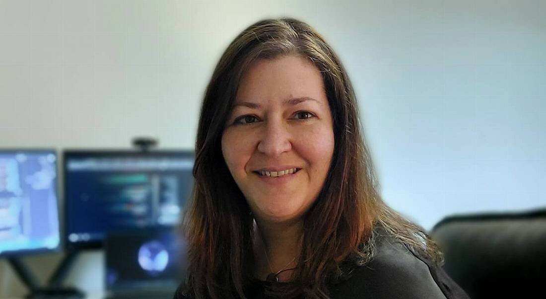 A woman smiles at the camera with a work desk and computer sitting behind her. She is Sumaya Almeida, a software engineer at Fidelity Investments.