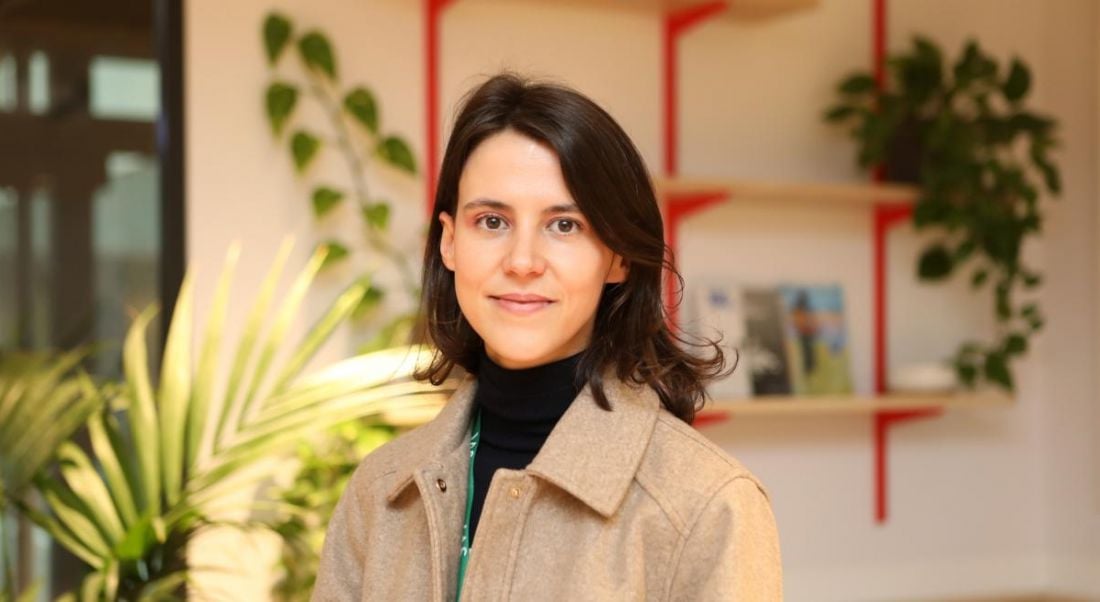 A woman wearing a beige jacket smiles at the camera in front of office plants and a shelf with books on it. She is Àngela García Martos, a senior associate technical account manager at Integral Ad Science.