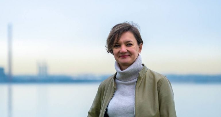A woman wearing a green jacket over a grey turtleneck smiles in front of a sea backdrop. She is Doctor Kasia Pluta, a quality assurance specialist at Amgen.