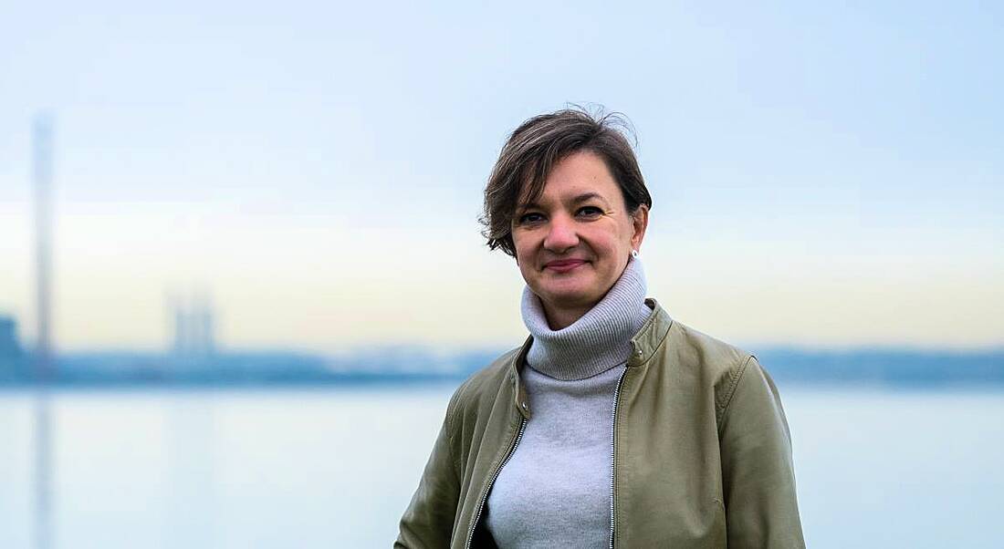 A woman wearing a green jacket over a grey turtleneck smiles in front of a sea backdrop. She is Doctor Kasia Pluta, a quality assurance specialist at Amgen.