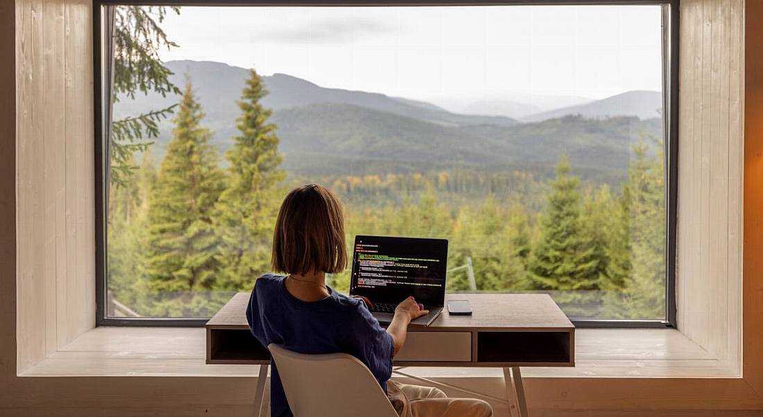 A woman is remote working at a desk, facing a scenic, rural view.