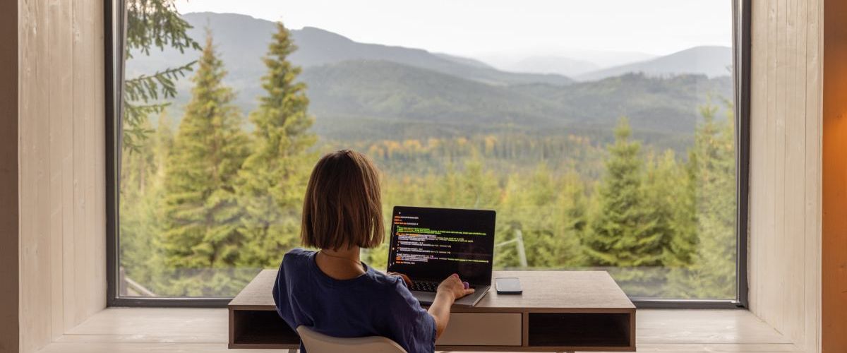 A woman is remote working at a desk, facing a scenic, rural view.