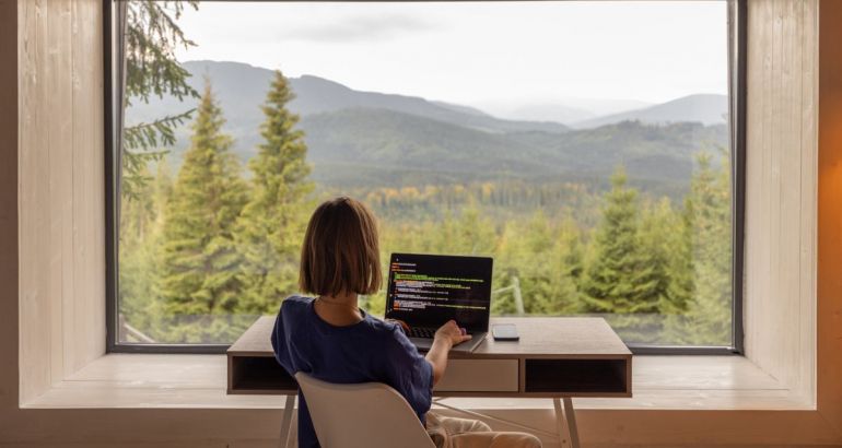A woman is remote working at a desk, facing a scenic, rural view.
