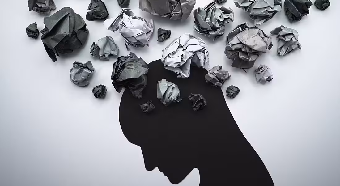 A figure in black looks down. Crumpled papers are expelled around the head, representing mental health in the workplace.