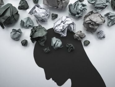 A figure in black looks down. Crumpled papers are expelled around the head, representing mental health in the workplace.
