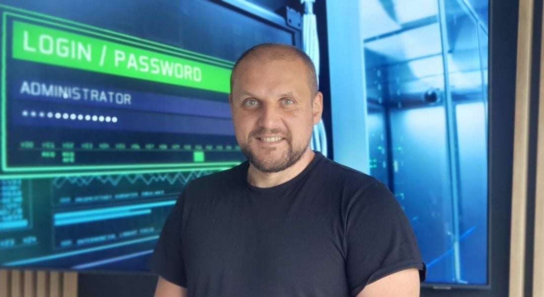 A man wearing a dark T-shirt smiles at the camera in front of a large computer monitor displaying computer login details. He is Damian Tumanowicz, a senior cybersecurity analyst at Liberty IT.