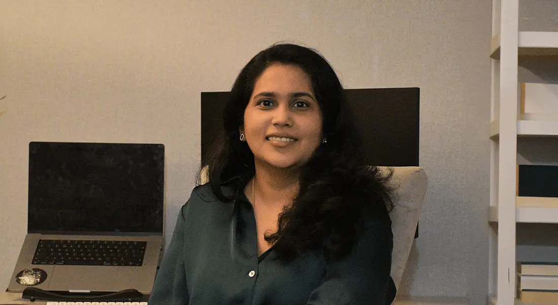 A woman wearing a dark green blouse sits smiling in front of a desk that holds a laptop and a monitor. She is Sanjana Mahadeshwar.