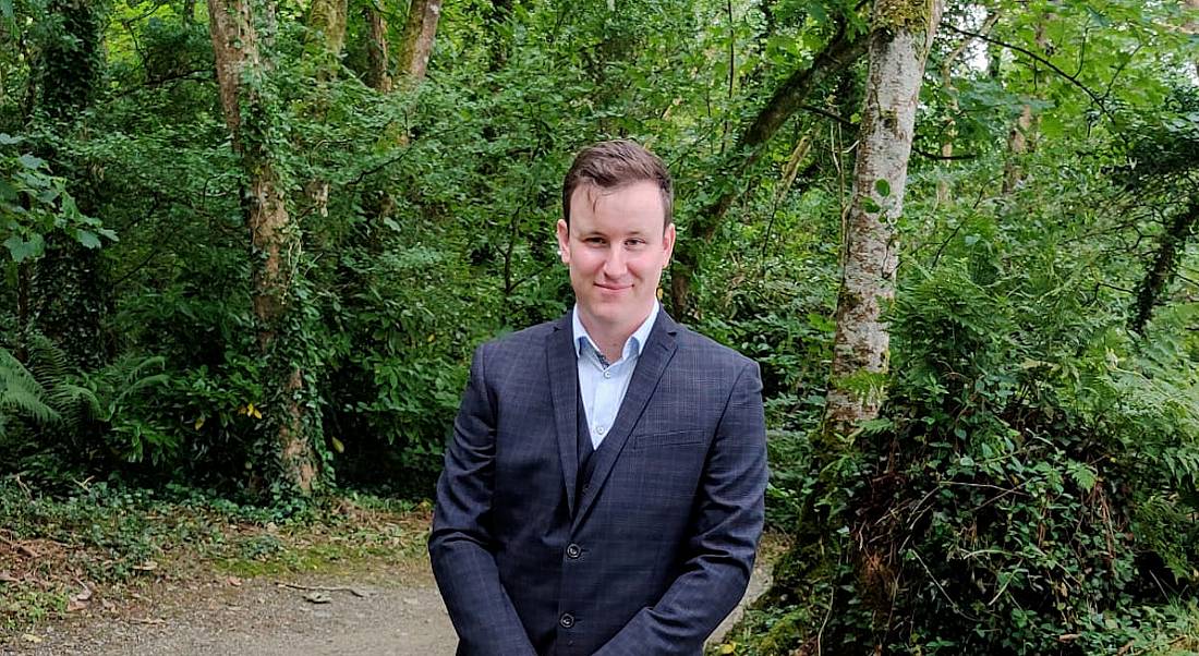 A man wearing a dark navy suit with a light blue open-collar shirt smiles in an outdoor setting. He is Sean Paterson, a senior technology engineer at Merck.