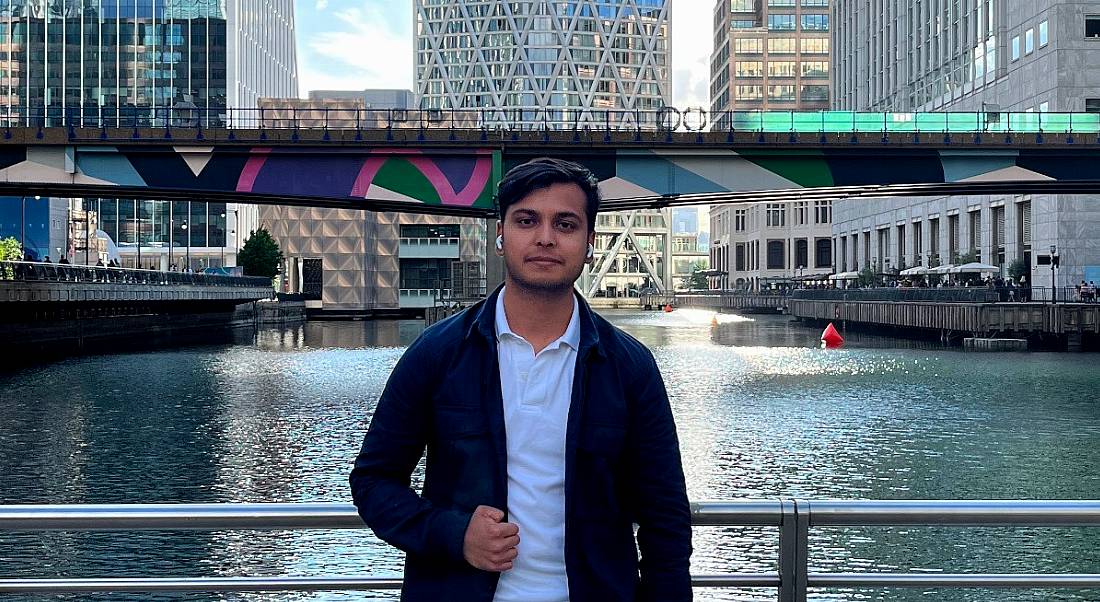 A man wearing a dark jacket over a blue shirt stands on a bridge overlooking a river, with another bridge seen behind him in a city landscape. He is Siddhant Goswami, a data engineer at Yahoo.